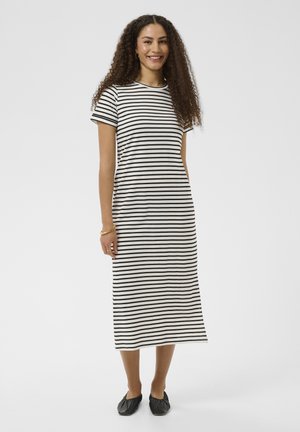 Kobieta z kręconymi włosami w czarno-białej pasiastiej sukience midi i czarnych balerinach, uśmiechająca się i stojąca na jednolitym tle.