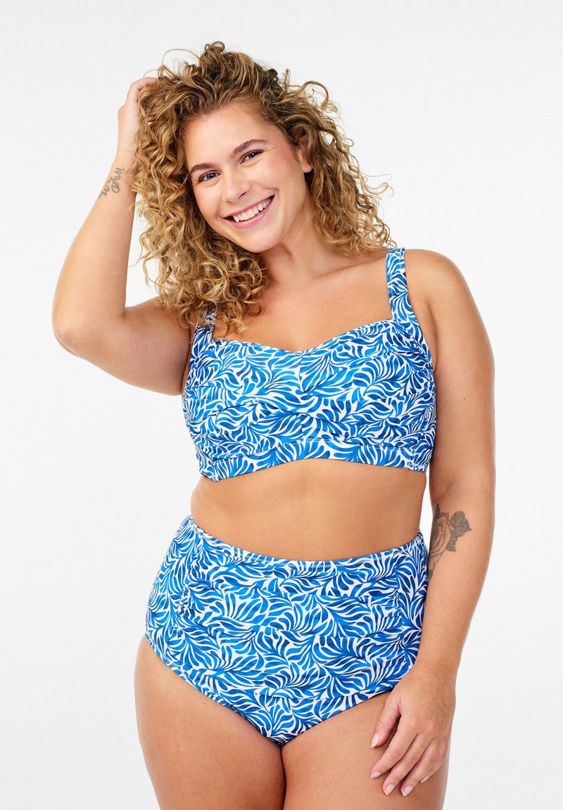 Smilende kvinde med krøllet hår iført blå og hvid mønstret to-delt badedragt, holder håret med den ene hånd, stående mod hvid baggrund.