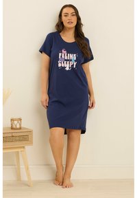 Kvinna som står barfota i en marinblå nattklänning med texten "Feline Sleepy" och färgglada kattillustrationer, bredvid ett litet träbord med ett ljus.