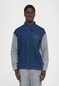 Homme portant une veste en denim bleue et grise avec "Fiorucci" brodé en rouge au-dessus d'une poche zippée sur la poitrine.