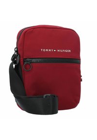 Borsa a tracolla rossa in nylon con tracolla nera, caratterizzata da una tasca frontale con zip e il logo "TOMMY HILFIGER" in bianco. Design rettangolare semplice.