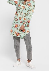 Tunique fleurie vert menthe avec des boutons, ornée de fleurs rouges et roses ainsi que de feuilles vertes. Associée à un pantalon gris texturé et des baskets blanches.