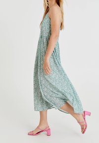 Vestido largo floral en verde con flores blancas, presenta tirantes finos y un dobladillo fluido. Combinado con tacones rosas con tiras.