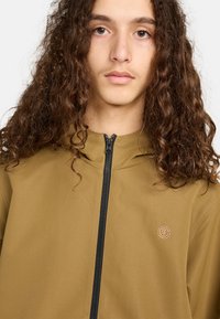 Junge Person mit langen, lockigen braunen Haaren, die eine olivgrüne Jacke mit Reißverschluss und einem kleinen runden Logo auf der Brust trägt.