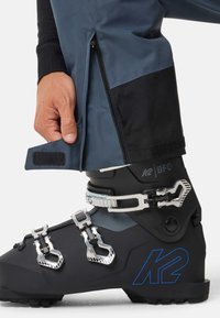 Schwarzer K2-Skiartikel mit drei metallischen Schnallen, einer glatten Oberfläche und einem blauen Logo. Winterhose mit Reißverschluss und Klettverschlussdetails ist sichtbar.