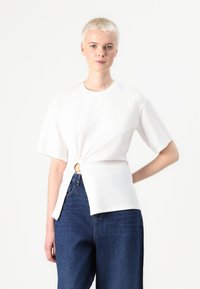 Tricou alb din bumbac cu mâneci scurte, având un detaliu cu nod pe lateral și un accent circular. Asortat cu blugi decolorati, largi, de culoare albastru închis.