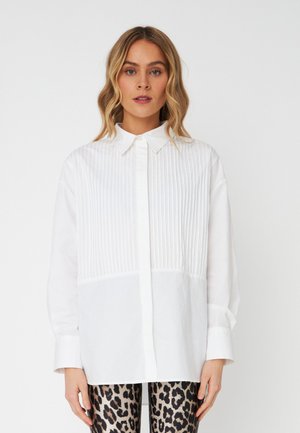 Camicia bianca con pieghe verticali sulla parte superiore davanti, colletto classico e chiusura con bottoni, abbinata a leggings con stampa animale a fantasia.