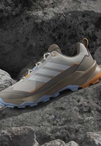 Chaussure de trail running marron clair et beige avec une semelle rugueuse et des bandes blanches, placée sur un terrain rocheux.