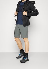 Abbigliamento outdoor da uomo con giacca nera con zip, t-shirt blu navy, pantaloni corti grigi e scarponi da trekking neri con dettagli blu.