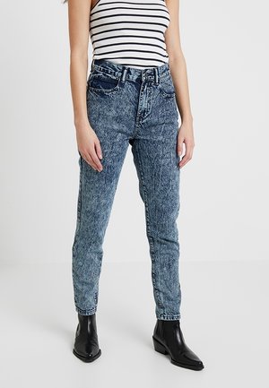 Kvinde iført højttaljede jeans med survask, sorte ankelstøvler og en hvid ærmeløs tanktop med sorte vandrette striber.