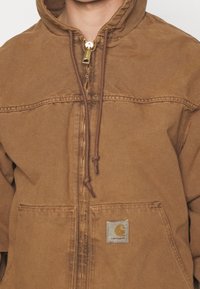 Brun zip-up jakke med hætte, lavet af slidstærkt stof. Har et logo patch, snoretræk detaljer og to forlommer.