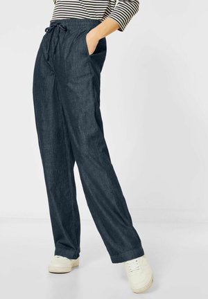 Persona con pantalones anchos azul oscuro con cintura de cordón, zapatillas blancas y camiseta de rayas blancas y negras, con las manos en los bolsillos.