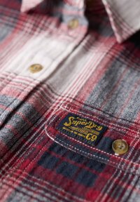 Chemise en flanelle à carreaux rouges et gris avec un tissu texturé, dotée d'une poche poitrine et d'un patch logo jaune et bleu, ainsi que de boutons marron.