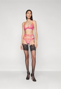 Lingerieset in felroze en oranje kant met een beha, jarretelband en bijpassende string. Voorzien van zwarte kanten kousen tot aan de dij.