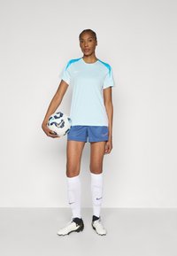 Αθλητική φανέλα Nike ανοιχτού μπλε με τυρκουάζ λεπτομέρειες, σε συνδυασμό με σκούρο μπλε σορτς με ροζ λογότυπο. Λευκές κάλτσες μέχρι το γόνατο και μαύρα παπούτσια ποδοσφαίρου.