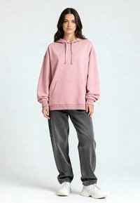 Felpa rosa in cotone con tasca frontale e coulisse regolabile, abbinata a jeans grigi larghi e sneaker bianche su uno sfondo neutro.
