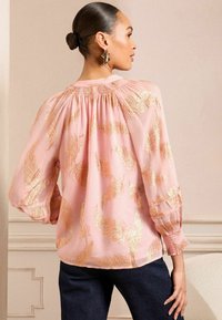 Rosa Bluse mit goldenen Blattmustern, gerafftem Ausschnitt und Ballonärmeln. Weiche Textur, kombiniert mit Jeans für den Stil.