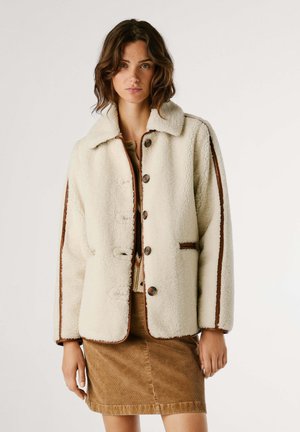 Veste en polaire crème avec des passepoils marron, cinq boutons et un col pointu. Superposée sur un haut beige et associée à une jupe en velours côtelé marron.