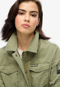 Olijfgroen jack met een kraag, knoopsluiting, twee borstzakken en een label met de tekst "SUPERDRY." De stof lijkt textuur en duurzaamheid te hebben.