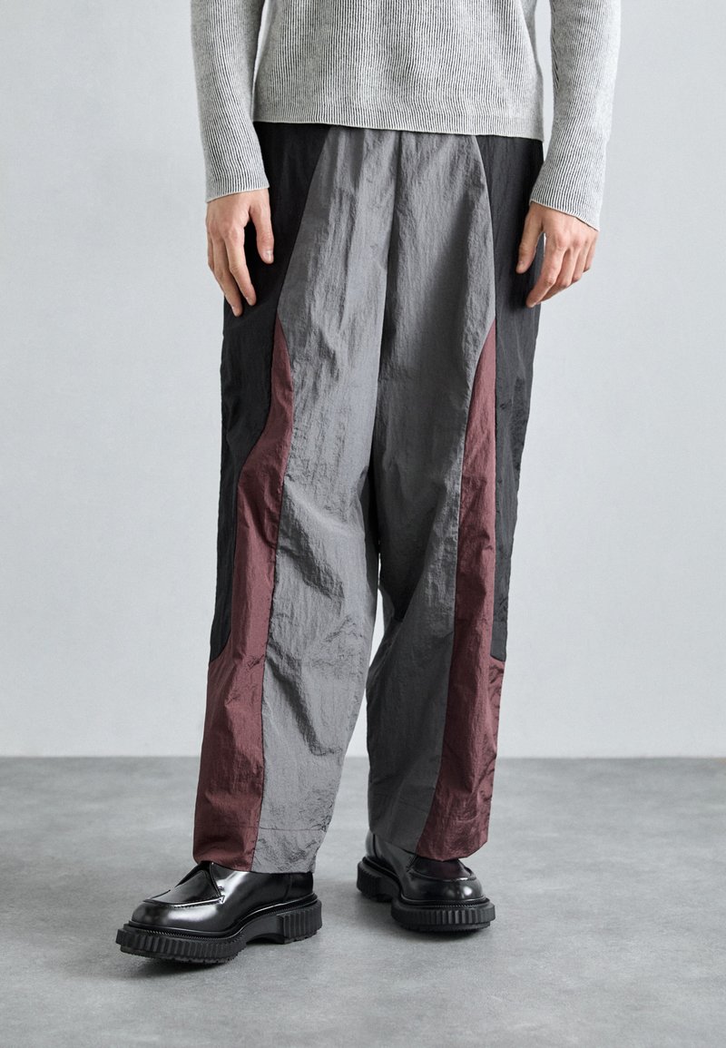 Pantalons larges gris et noirs avec des panneaux latéraux bordeaux, fabriqués en tissu léger et texturé. Portés avec des chaussures noires brillantes.