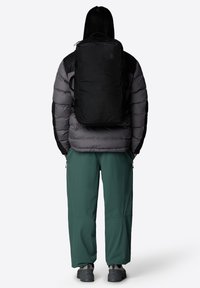 Svart ryggsäck med en modern design, tillverkad av tåligt material, bärs över en grå pufferjacka, kombinerad med lösa gröna byxor och grå skor.