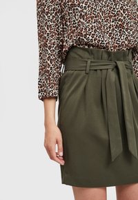 Jupe vert olive avec une ceinture en tissu nouée, présentant des plis et une texture lisse. Associée à un chemisier à manches longues imprimé léopard.