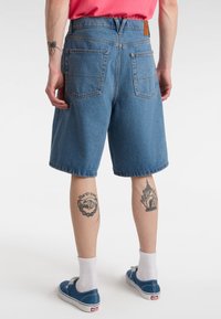 Niebieskie szorty z denimu z wysokim stanem, wyposażone w dwie tylne kieszenie i skórzaną naszywkę. Noszone z niebieskimi trampkami i białymi skarpetkami.