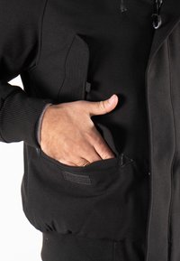 Veste noire avec poignets côtelés, fermeture éclair à l'avant et plusieurs poches, dont une avec fermeture Velcro. Texture lisse.