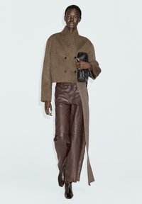 Donna che indossa un giubbotto marrone testurizzato doppiopetto, pantaloni in pelle con pannello in maglia, clutch nera intrecciata e stivali neri con tacco, che cammina in avanti.
