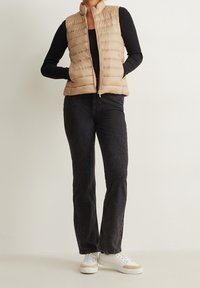 Beżowa kamizelka puchowa z wysokim kołnierzem, zamkiem z przodu i pikowaną fakturą, w zestawieniu z czarnymi spodniami typu straight-leg i białymi trampkami.