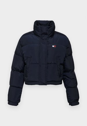 Apgriezta, tumši zila pūkaina jaka ar augstu apkakli, quilted tekstūru un logo detaļu kreisajā krūtī. Tam ir garas piedurknes un priekšējais rāvējslēdzis.