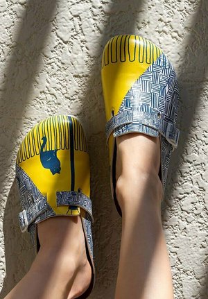 Feet wearing yellow and white patterned slip-on shoes with blue cat and stripe designs on textured light wall background.