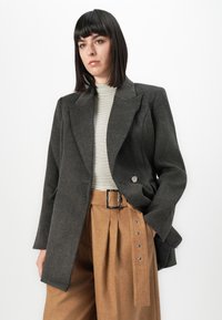 Femme avec des cheveux noirs lisses portant un blazer surdimensionné gris foncé, un col roulé rayé et un pantalon marron taille haute avec une ceinture.