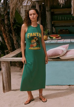 Woman with curly hair stands barefoot in a green sleeveless dress with "Fanta Orange" print near a tropical bar with fruit and cushions.