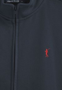 Giacca a zip blu navy realizzata in tessuto morbido, con colletto alto e un piccolo logo ricamato di un golfista rosso sul lato sinistro del petto.