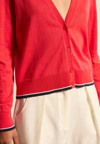 Roter Cardigan mit tiefem V-Ausschnitt, fünf Knöpfen und gerippten Bündchen. Verziert mit marinen und weißen gestreiften Akzenten am Saum und den Bündchen. Weicher Stoff.