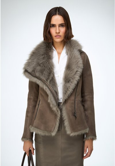 Veste en cuir marron avec col en fausse fourrure grise, fermeture éclair à l'avant et deux poches latérales. Portée sur une chemise blanche, assortie à une jupe.