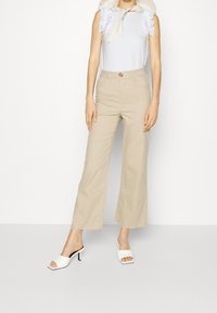 Beżowe spodnie o szerokich nogawkach z wysokim stanem, zapinane na guziki, uszyte z miękkiego materiału, w połączeniu z białą bluzką bez rękawów ozdobioną falbankami.