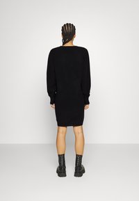 Robe pull noire en maille avec manches longues, coupe ample, ourlet côtelé et détail à l'encolure arrière. Associée à des bottines noires épaisses.
