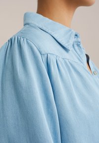 Blaue Denimbluse mit weicher Textur, Spitzkragen und Falten detaillierte Schultern. Seitenknopfverschluss sichtbar.