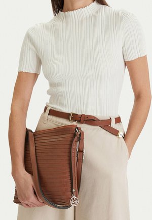 Mujer con una camiseta blanca de manga corta acanalada y pantalones beige con cinturón marrón, sosteniendo un gran bolso de hombro de cuero marrón.