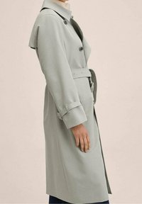 Jasnoszara trenchcoat z paskiem w talii, guzikami z przodu oraz wyraźnym kołnierzem. Posiada paski na mankietach oraz gładką fakturę materiału.