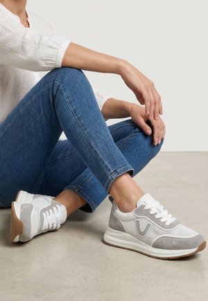 Osoba sedící na podlaze se zkříženýma nohama, oblečená v modrých džínách, bílých a šedých teniskách a bílém tričku s dlouhým rukávem.