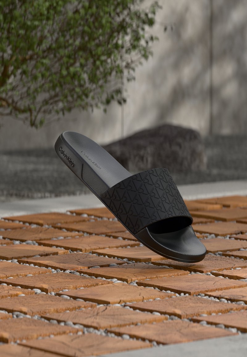 Sandale mule noire Calvin Klein avec lanière embossée reposant sur une surface carrelée extérieure, avec un arrière-plan de verdure et de pierres.