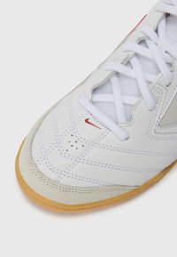 Weißer Sportschuh mit einem strukturierten Lederobermaterial, Mesh-Akzenten, rotem Logo und einer Sohle aus Gummi. Weiße Schnürsenkel sorgen für einen sicheren Sitz.