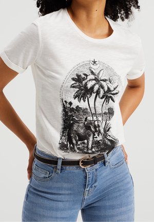 Kobieta w białym t-shircie z czarną vintage’ową ilustracją słonia i palmy, w połączeniu z niebieskimi jeansami i czarnym paskiem ze złotą klamrą.