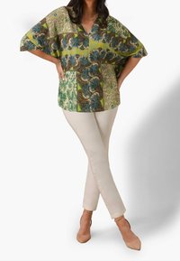 Blusa colorata e stampata con scollatura a V, maniche corte e design fluido; abbinata a pantaloni attillati beige chiaro e tacchi nudi.
