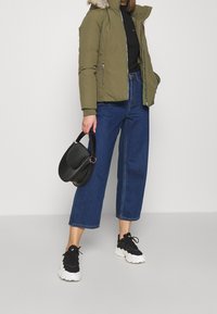 Olivgrön puffjacka med avtagbar pälsluva, kombinerad med mörkblå vida jeans och svarta sportskor. Håller en svart handväska.