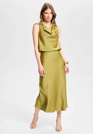 Femme debout portant une robe sans manches en satin vert olive avec un col bénitier, un bracelet en or et des sandales à talons hauts dorées à brides.