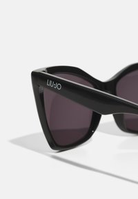 Occhiali da sole a forma di occhio di gatto neri con lenti scure. Presentano una texture prominente sulla montatura e il logo "LIU·JO" sulla stanghetta.